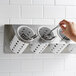 A hand holding a spoon in a Steril-Sil stainless steel flatware organizer with white plastic cylinders.