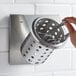 A hand placing a spoon in a Steril-Sil stainless steel flatware organizer with a perforated metal cylinder.