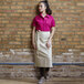 A woman wearing a Uncommon Chef sand beige canvas apron with natural webbing standing against a brick wall.