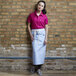 A woman wearing a sky blue Uncommon Chef bistro apron with natural webbing standing in front of a brick wall.