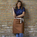 A woman in a blue shirt and Uncommon Chef brown canvas waist apron with natural webbing leaning against a brick wall.