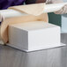 A person in gloves using a white foam square cake dummy to cut a cake.