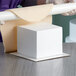A person rolling a white Baker's Mark foam square cake dummy on a table.