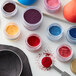 A jar of Roxy & Rich Neon Pink Fondust powder on a white surface with other colorful jars of powder.