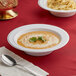 A white plastic bowl of soup with a spoon on a red tablecloth.