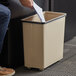 A person's hand putting paper into a beige Rubbermaid wastebasket.