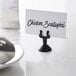 An American Metalcraft black metal low profile harp card holder on a counter with a place card in it.