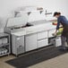 A woman using an Avantco refrigerated pizza prep table in a large commercial kitchen.