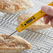 A hand using a yellow Comark digital pocket probe thermometer to check the temperature of meat on a grill.