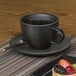 A Front of the House semi-matte black porcelain saucer with a cup of tea and a plate of fruit.