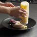 A person pouring a yellow sauce onto a Front of the House Spiral Ink semi-matte black porcelain plate with food.