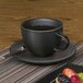 A close up of a black Front of the House Spiral Ink porcelain cup with liquid in it and a plate of fruit.