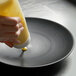 A hand pouring yellow mustard onto a Front of the House Spiral Ink black porcelain plate.