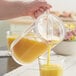 A hand pouring orange juice from a Carlisle Elan clear pitcher into a glass.