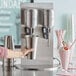 A person holding a silver cup of a pink milkshake in front of an AvaMix double spindle milkshake machine.