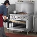 A man in a chef's uniform using a Cooking Performance Group liquid propane range to put food in an oven.