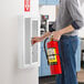 A man holding a JL Industries Ambassador series white steel fire extinguisher cabinet with a full window recessed in a wall.