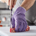 A person cutting meat with a knife while wearing purple San Jamar cut resistant gloves.
