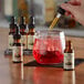 A hand using a dropper to add King Floyd's Bitters to a glass of red liquid with ice.