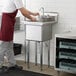 A man in a red apron washing dishes in a Steelton stainless steel commercial sink.