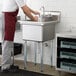 A man in a red apron washing a glass in a Steelton one compartment sink.