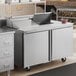 A stainless steel two-door refrigerated sandwich prep table with a raised ingredient rail and casters.