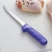 A Dexter-Russell purple narrow boning knife with a blue handle on a white surface next to a piece of chicken.