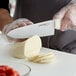 A person using a Dexter-Russell scalloped chef knife to cut a piece of cheese on a table.