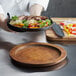 A person holding a pan of food on a Valor round rubberwood underliner with food on a plate.