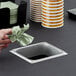 A hand dropping a green napkin into a rectangular stainless steel in-counter trash chute.