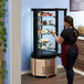 A woman wearing a red apron standing in front of an Avantco copper circular glass refrigerated display case filled with cakes.