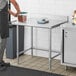 A man in an apron standing next to a Regency stainless steel open base work table.