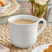 An Acopa Capri white stoneware cup filled with a brown liquid on a table.