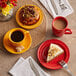 A red Acopa Capri stoneware mug filled with a liquid on a table with food and coffee.