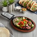 A Valor oval cast iron fajita skillet with meat and vegetables next to a plate of tacos on a wood surface.