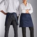 A man and woman wearing navy blue half bistro aprons with pockets.