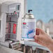 A hand holding a Rubbermaid Microburst 9000 Linen Fresh air freshener spray can near a dispenser.