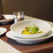 A white oval slanted porcelain bowl filled with salad on a white plate on a table.