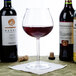 A customizable Tritan plastic balloon wine glass filled with red wine on a table next to bottles of wine.