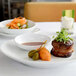 A white oval Front of the House ramekin filled with brown liquid on a table with a plate of bacon wrapped steak and greens.