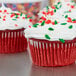 A close-up of a red cupcake in a white Hoffmaster fluted wrapper with white frosting and sprinkles.