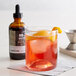 A glass of whiskey with ice and an orange slice next to a bottle of 18.21 Bitters.