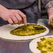 A person using a spatula to spread green sauce on a Rich's Fresh N Ready pizza crust on a counter.