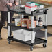 A woman pushing a black utility cart with food containers on the shelves.