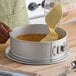 A round anodized aluminum springform cake pan with a latch, being filled with batter over a graham cracker crust.