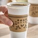 A white disposable coffee cup with a brown printed sleeve featuring coffee bean and leaf designs.