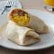 A plate with a fork and a Father Sam's Bakery flour tortilla, a burrito with scrambled eggs and meat, and a glass of orange juice.