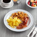 An Acopa Foundations white melamine plate with breakfast food and a cup of coffee on a table with a fork and napkin.