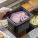 A gloved hand holding a Vigor 1/6 size black polycarbonate food pan of sliced onions.