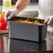 A person in gloves holding a black Vigor polycarbonate food pan filled with vegetables.